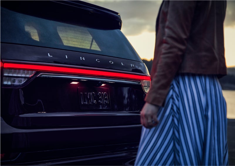 A person is shown near the rear of a 2023 Lincoln Aviator® SUV as the Lincoln Embrace illuminates the rear lights | Kirk Brothers Lincoln of Vicksburg in Vicksburg MS