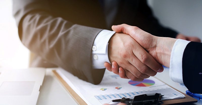 Two business professionals shaking hands over a desk with charts and a laptop.
